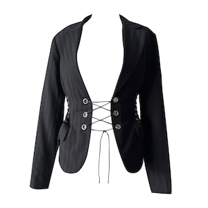 Puede incluir: Un blazer negro de rayas diplomáticas con mangas largas, solapa de muesca y un cierre frontal que consiste en tres pares de ojales metálicos plateados entrelazados con un cordón negro fino que cuelga en la cintura, creando un efecto de corsé.