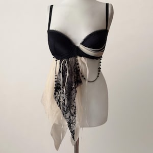 Puede incluir: Un sujetador negro con diseño de copas estructuradas y detalles en tela blanca. La prenda presenta una superposición asimétrica y drapeada de tela color crema transparente y encaje negro, adornada con una delicada cadena de pequeñas cuentas negras.