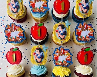 May include: A set of twelve cupcakes decorated with Snow White themed toppers, including images of Snow White in a gold frame, red apples with the number one, and groups of seven dwarfs. The cupcakes feature various colored frosting.
