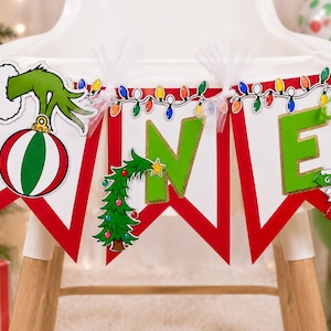 May include: A decorative banner spelling the word ONE with white pennants featuring red borders. The letter O is replaced by a green hand holding a striped ornament, the N by a decorated Christmas tree, and the E by a signpost reading Grinch's Lair, Whoville, and Mount Crumpit.