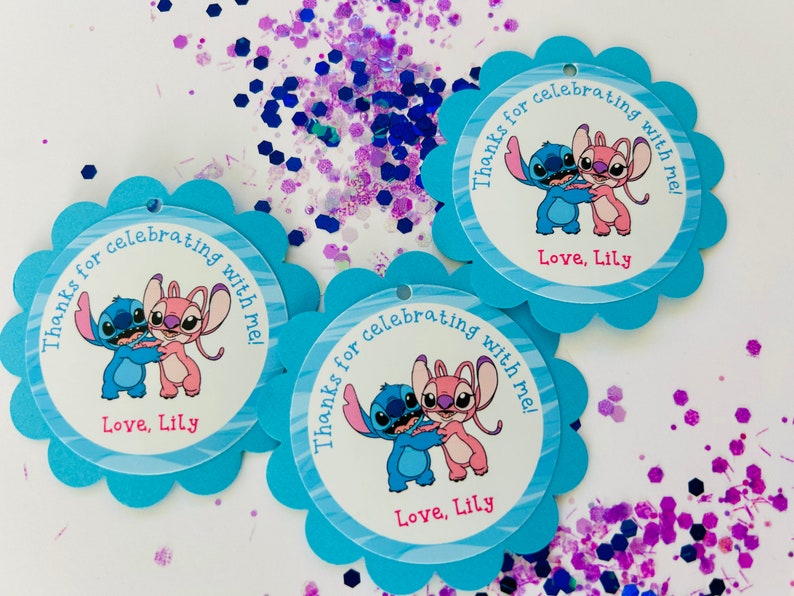 K&ouml;nnte beinhalten: Drei blaue und wei&szlig;e Geschenkanh&auml;nger mit gewelltem Rand. Jeder Anh&auml;nger zeigt ein Cartoon-Bild von Stitch und Angel aus dem Disney-Film Lilo & Stitch. Der Text auf den Anh&auml;ngern lautet "Danke, dass du mit mir gefeiert hast! Liebe, Lily."