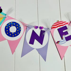 May include: A pink and purple banner with the word "ONE" in large purple letters. The banner is decorated with Minnie Mouse characters, a life preserver, a beach umbrella, and a unicorn float.