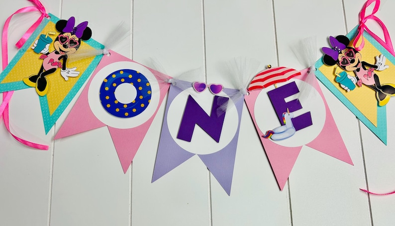 May include: A pink and purple banner with the word "ONE" in large purple letters. The banner is decorated with Minnie Mouse characters, a life preserver, a beach umbrella, and a unicorn float.