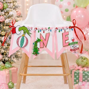 May include: A high chair banner featuring the word ONE in pink letters. The letter O is replaced by a Grinch hand holding a striped ornament, the N is a Christmas tree, and the E features a signpost with Grinch's Lair, Whoville, and Mount Crumpit.