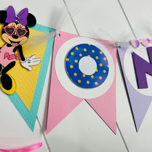 May include: A colorful banner with a Minnie Mouse character holding a drink, a blue and yellow life preserver, and the letter "N" in purple. The banner is decorated with white tulle and has a pink, yellow, and blue color scheme.