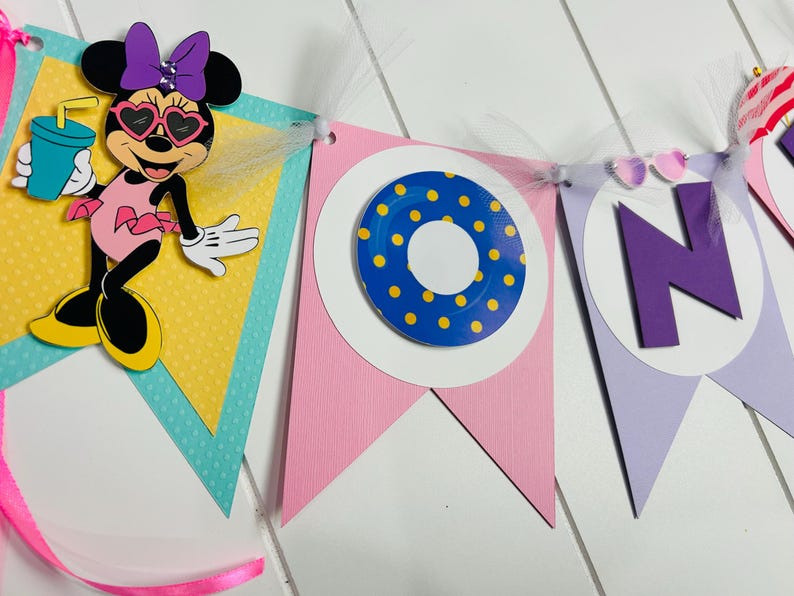 May include: A colorful banner with a Minnie Mouse character holding a drink, a blue and yellow life preserver, and the letter "N" in purple. The banner is decorated with white tulle and has a pink, yellow, and blue color scheme.