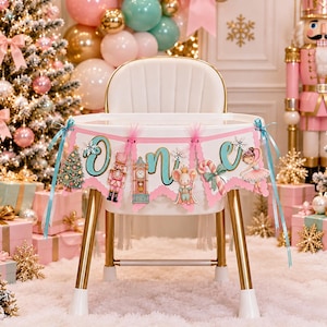 May include: A white high chair with gold legs features a pink and teal banner spelling out the word ONE. The banner is decorated with illustrations of a Christmas tree, a nutcracker, a clock, a mouse, a candy cane, and a ballerina.