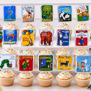 May include: A collection of fifteen cupcakes topped with miniature rectangular signs featuring classic children's book covers. The signs are arranged in three rows on a white stand, surrounded by stacks of the corresponding books on a white surface.