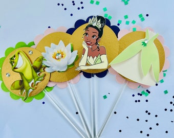 May include: Four cupcake toppers featuring characters from the Disney movie The Princess and the Frog. The toppers are on sticks and feature Tiana, a frog, a lily pad, and a green dress.