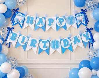 May include: A two-tier Happy Birthday banner featuring blue and white pennants with beach-themed accents like seashells and umbrellas, decorated with blue ribbons and surrounded by a balloon arch of blue, white, and clear confetti-filled balloons.