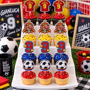 May include: A tiered display of twelve cupcakes topped with soccer-themed decorations including red jerseys with the number 9, Spanish crests, blue number 9s with stars, and soccer balls, all featuring colorful sprinkles on the frosting.