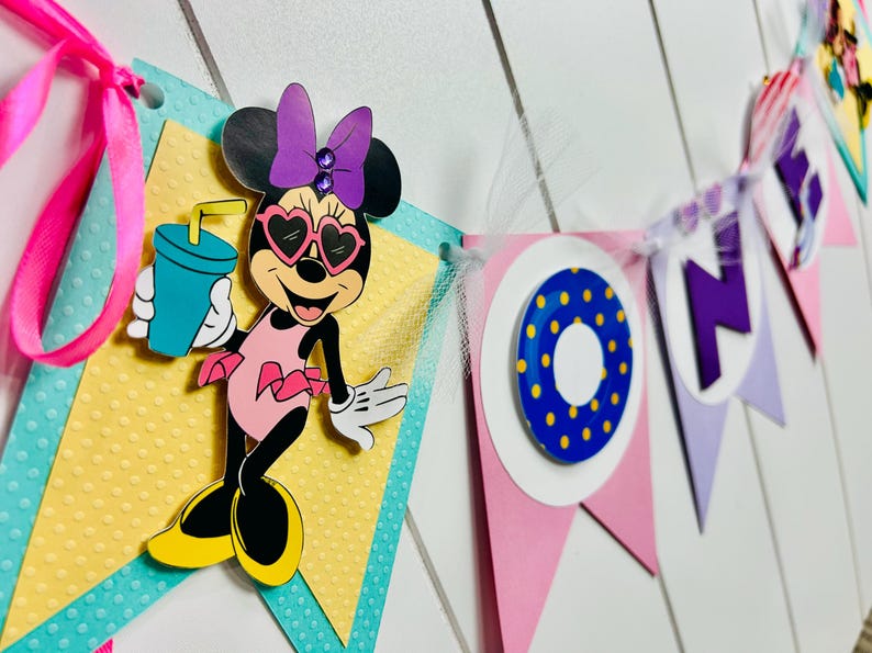 May include: A pink, purple, and blue banner with the word "ONE" in purple letters. The banner features a Minnie Mouse character wearing sunglasses and a pink swimsuit, holding a blue drink with a straw. The banner is decorated with yellow polka dots and a blue and yellow ring.