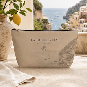 May include: A beige zippered pouch featuring the text LA DOLCE VITA. POSITANO . IT, a small lemon branch illustration, and a faded graphic of a coastal town. The pouch has a black zipper closure and a textured fabric finish on the exterior surface.