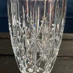 May include: A clear glass vase with a starburst and vertical line pattern. The vase has a wide opening and a slightly tapered shape. The glass is thick and has a reflective quality, catching the light and creating a sparkling effect.