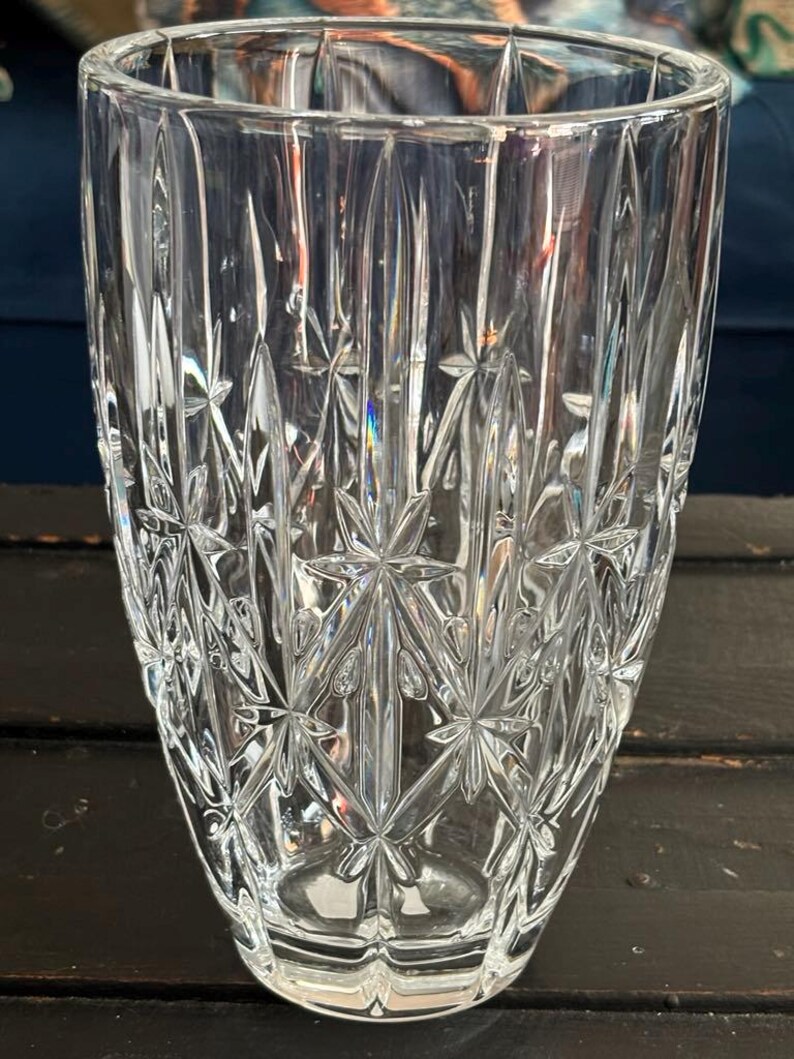 May include: A clear glass vase with a starburst and vertical line pattern. The vase has a wide opening and a slightly tapered shape. The glass is thick and has a reflective quality, catching the light and creating a sparkling effect.