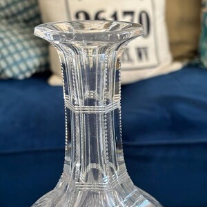 May include: A clear, cut-glass decanter with a flared lip and a faceted body. The decanter features intricate geometric patterns and a long, slender neck. The background includes a blue surface and a pillow with text.