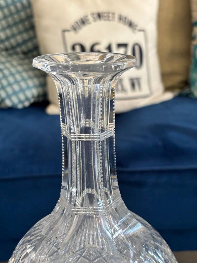 May include: A clear, cut-glass decanter with a flared lip and a faceted body. The decanter features intricate geometric patterns and a long, slender neck. The background includes a blue surface and a pillow with text.