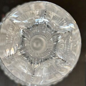 May include: Clear crystal glass with intricate starburst and geometric patterns. The glass has a circular base with a detailed design, reflecting light and creating a sparkling effect. The glass is likely part of a glassware set.