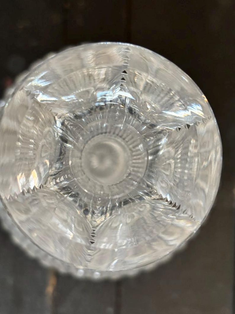 May include: Clear crystal glass with intricate starburst and geometric patterns. The glass has a circular base with a detailed design, reflecting light and creating a sparkling effect. The glass is likely part of a glassware set.