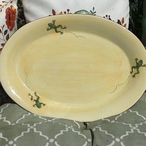 May include: Oval, cream-colored ceramic platter with three green frog illustrations. The frogs appear to be jumping, with curved lines indicating their path. The platter has a slightly raised edge and is set against a background of pillows.