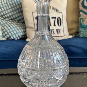 May include: A clear, cut-glass decanter with a bulbous body and a flared neck. The decanter features intricate geometric patterns and vertical lines, reflecting light. The base has a starburst design. A decorative piece for serving beverages.