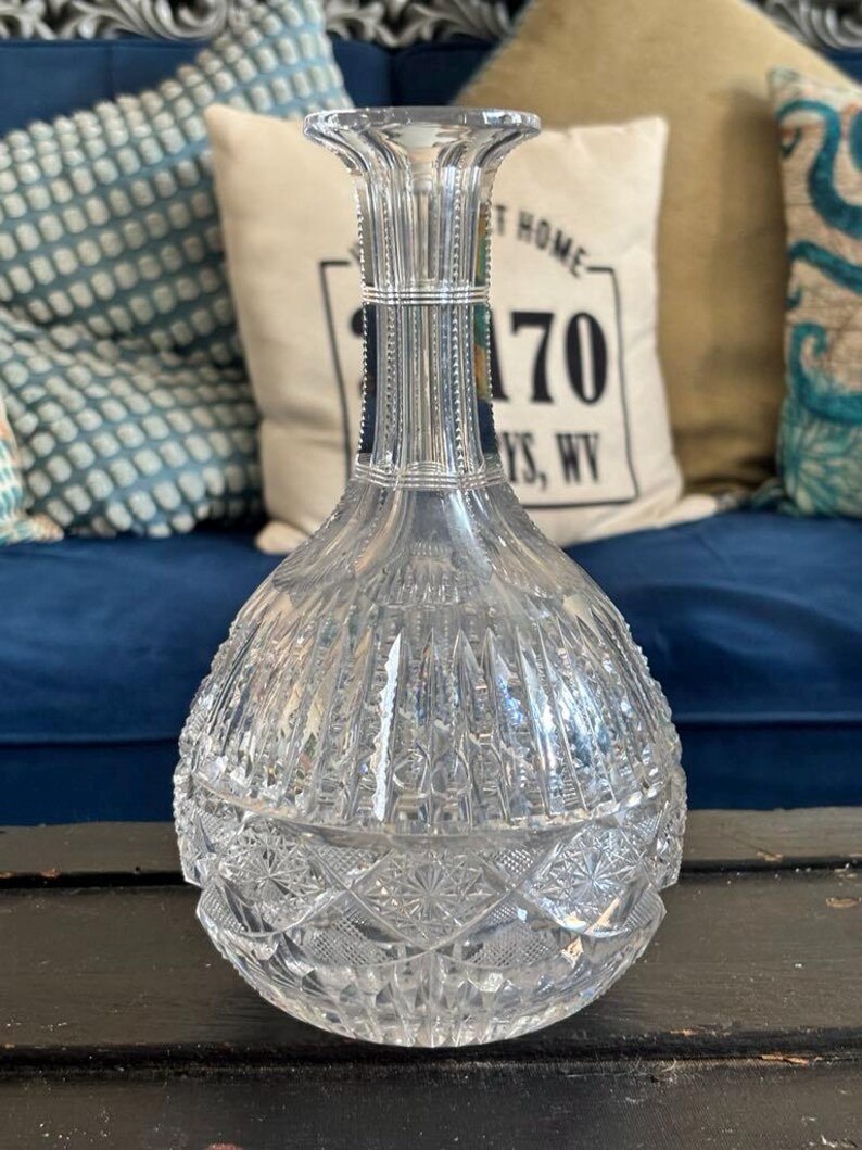 May include: A clear, cut-glass decanter with a bulbous body and a flared neck. The decanter features intricate geometric patterns and vertical lines, reflecting light. The base has a starburst design. A decorative piece for serving beverages.