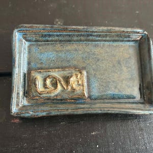 May include: A rectangular ceramic tray with a glossy blue and brown glaze. The tray features a raised rectangular section on the left side with the word LOVE embossed in a textured, metallic-toned finish, showing uneven edges.