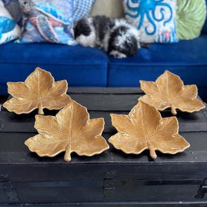 May include: Four ceramic leaf-shaped serving dishes in a warm, golden-brown hue. The dishes are arranged on a dark, wooden trunk. The background features a blue sofa with decorative pillows.
