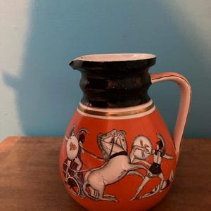 May include: A small, orange ceramic pitcher with a black and gold rim and handle. The pitcher features a classical scene of figures, horses, and a chariot. The background is a light blue.