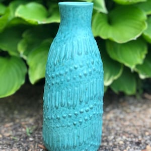 May include: A turquoise ceramic vase with a textured surface. The vase has a narrow neck and a rounded body with vertical lines and small, evenly spaced dots. The vase is approximately 8 inches tall.