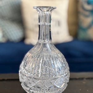 May include: A clear, cut-glass decanter with a bulbous body and a flared neck. The decanter features intricate geometric patterns and vertical lines, reflecting light. The base has a detailed starburst design. A decorative piece for serving beverages.