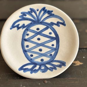 May include: A small, round, off-white ceramic plate with a hand-painted blue pineapple design. The pineapple features a cross-hatched pattern and dot details. The plate is likely for decorative or serving purposes.