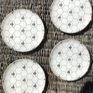 May include: Four white ceramic plates with a honeycomb pattern and bee illustrations. Each plate has a gold rim. The plates are arranged on a woven wicker surface.