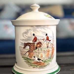 May include: A white ceramic jar with a lid, featuring a hunting scene with riders on horseback, a dog, and a wooden fence. The jar has a green trim on the base and lid, and the lid has a small knob.