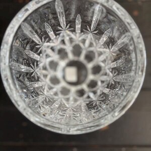 May include: A clear cut-crystal glass with a detailed starburst and geometric pattern. The glass has a thick base and a wide opening, showcasing a detailed design. The cut crystal reflects light, creating a sparkling effect. Likely a decorative item.