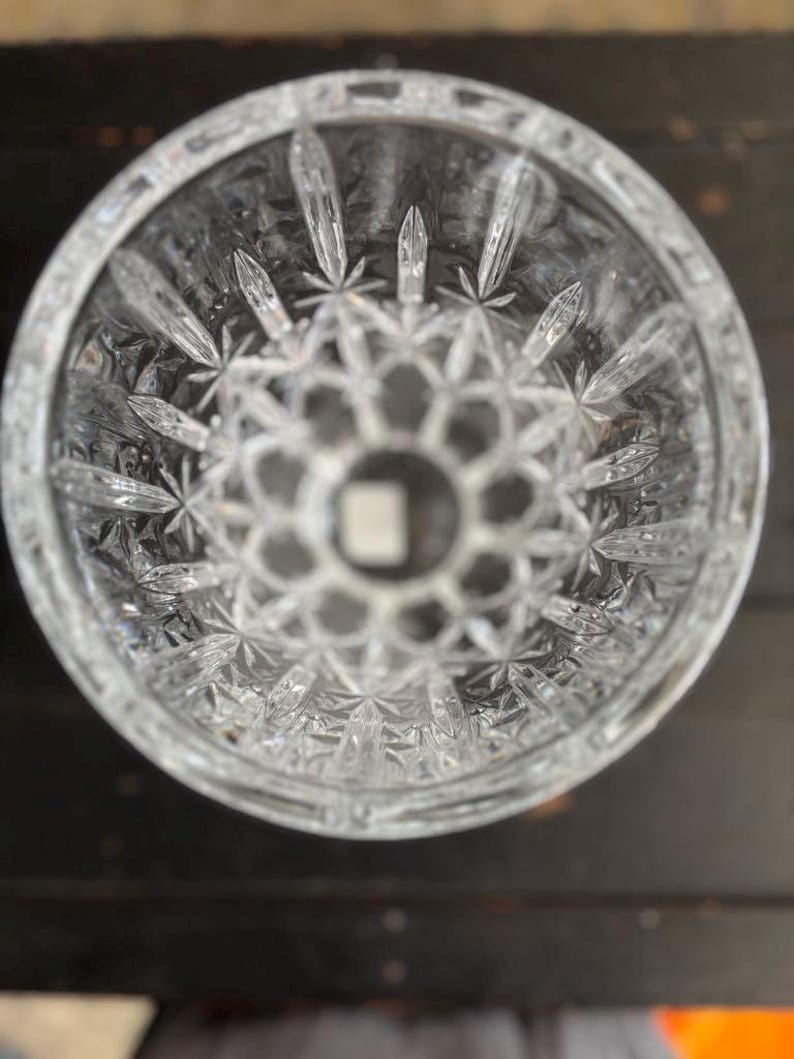 May include: A clear cut-crystal glass with a detailed starburst and geometric pattern. The glass has a thick base and a wide opening, showcasing a detailed design. The cut crystal reflects light, creating a sparkling effect. Likely a decorative item.
