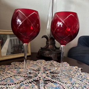 May include: Two ruby red wine glasses with a diamond pattern etched into the bowl. The glasses have long, clear stems and sit on a decorative, colorful doily. The background includes a lamp and a framed picture.