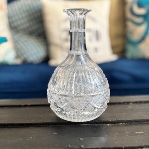 May include: A clear, cut-glass decanter with a bulbous base and a slender neck. The decanter features intricate geometric patterns and vertical lines, reflecting light. The background includes blue and white pillows, suggesting a decorative setting.
