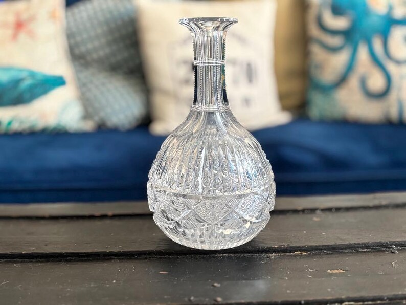 May include: A clear, cut-glass decanter with a bulbous base and a slender neck. The decanter features intricate geometric patterns and vertical lines, reflecting light. The background includes blue and white pillows, suggesting a decorative setting.