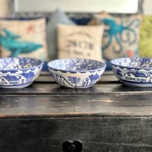 May include: Three blue and white ceramic bowls with a speckled pattern and a house design around the rim. The bowls are arranged on a dark wooden surface, with decorative pillows in the background.