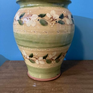 May include: A handcrafted ceramic vase with a wide, rounded body and a slightly flared rim. The vase features a cream-colored base with green and tan horizontal bands. White floral designs with green leaves and stems decorate the vase.