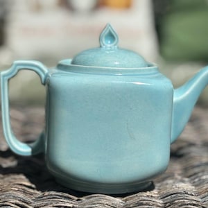 May include: A light blue ceramic teapot with a matching lid. The teapot has a curved spout and a handle with a decorative design. The lid is topped with a teardrop-shaped finial. The teapot is sitting on a textured surface.