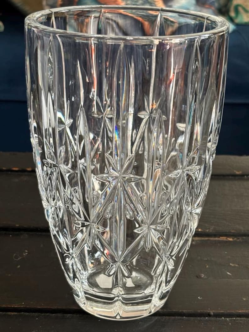 May include: A clear, cut-glass vase featuring a starburst and vertical line design. The vase has a cylindrical shape with a gently widening top. The detailed pattern reflects light, producing a shimmering effect. The vase is empty and rests on a dark surface.