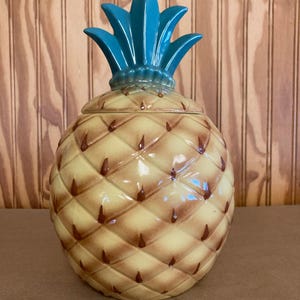 May include: A ceramic pineapple-shaped jar with a lid. The jar is primarily yellow with brown diamond-shaped patterns and brown accents. The top of the pineapple is a vibrant teal color.