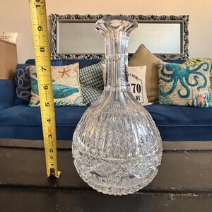 May include: A clear, cut-glass decanter with a bulbous base and a tall, fluted neck. The decanter is approximately 10 inches tall. The intricate cut-glass design features vertical and geometric patterns, reflecting light.