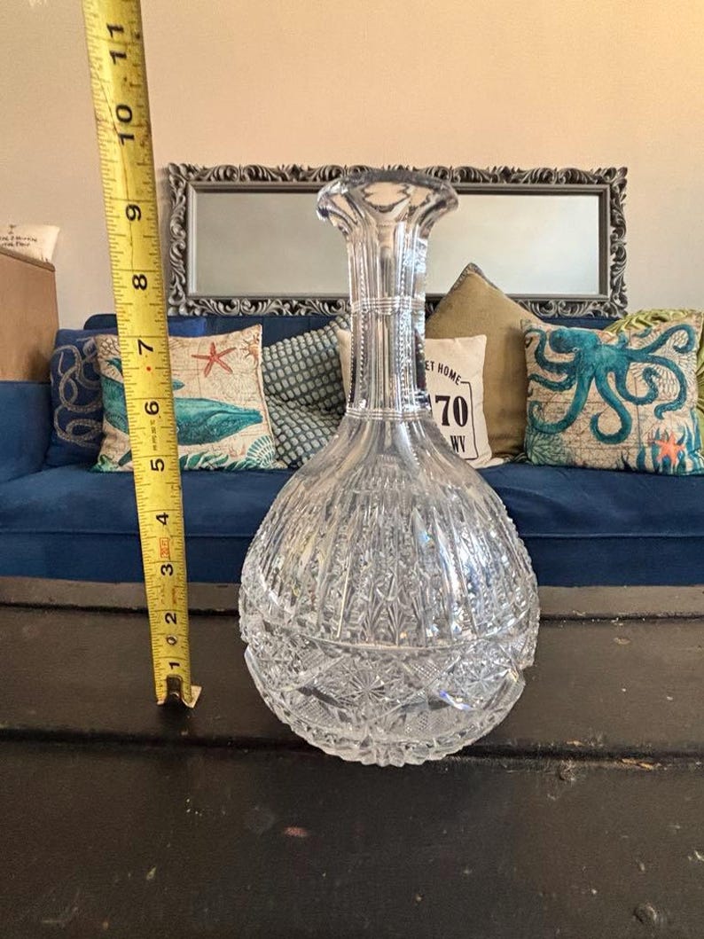May include: A clear, cut-glass decanter with a bulbous base and a tall, fluted neck. The decanter is approximately 10 inches tall. The intricate cut-glass design features vertical and geometric patterns, reflecting light.