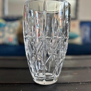 May include: A clear, cut-glass vase with a starburst pattern. The vase has a wide, cylindrical shape with vertical cuts and a detailed, geometric design. The glass reflects light, creating a sparkling effect. The vase is sitting on a dark wooden surface.