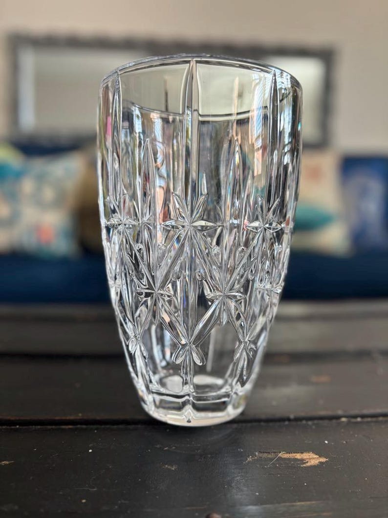 May include: A clear, cut-glass vase with a starburst pattern. The vase has a wide, cylindrical shape with vertical cuts and a detailed, geometric design. The glass reflects light, creating a sparkling effect. The vase is sitting on a dark wooden surface.