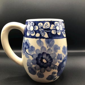 May include: A ceramic mug with a white handle and a rounded body. The mug is decorated with a blue floral pattern against a white background. The top of the mug features a band of blue and white floral designs.