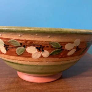 May include: A hand-painted ceramic bowl with a floral design. The bowl features a green rim, a band of orange and brown stripes, and a pink base. White flowers and green leaves are painted around the bowl.